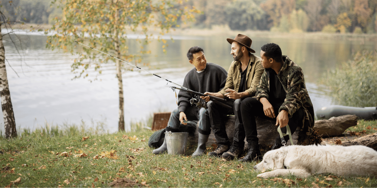 Three men in sitting on a log and talking near a lake with one holding and rod and reel