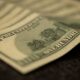 Minimalist photo of a single dollar bill caught on a sharp fish hook against a clean background, symbolizing the high cost...